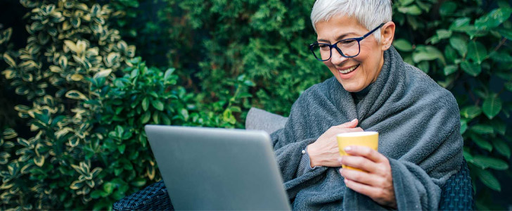 Older woman wrapped in blanket outside smiling at laptop screen