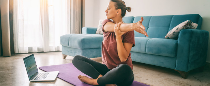 Woman sitting on yoga mat in living room stretching out arm